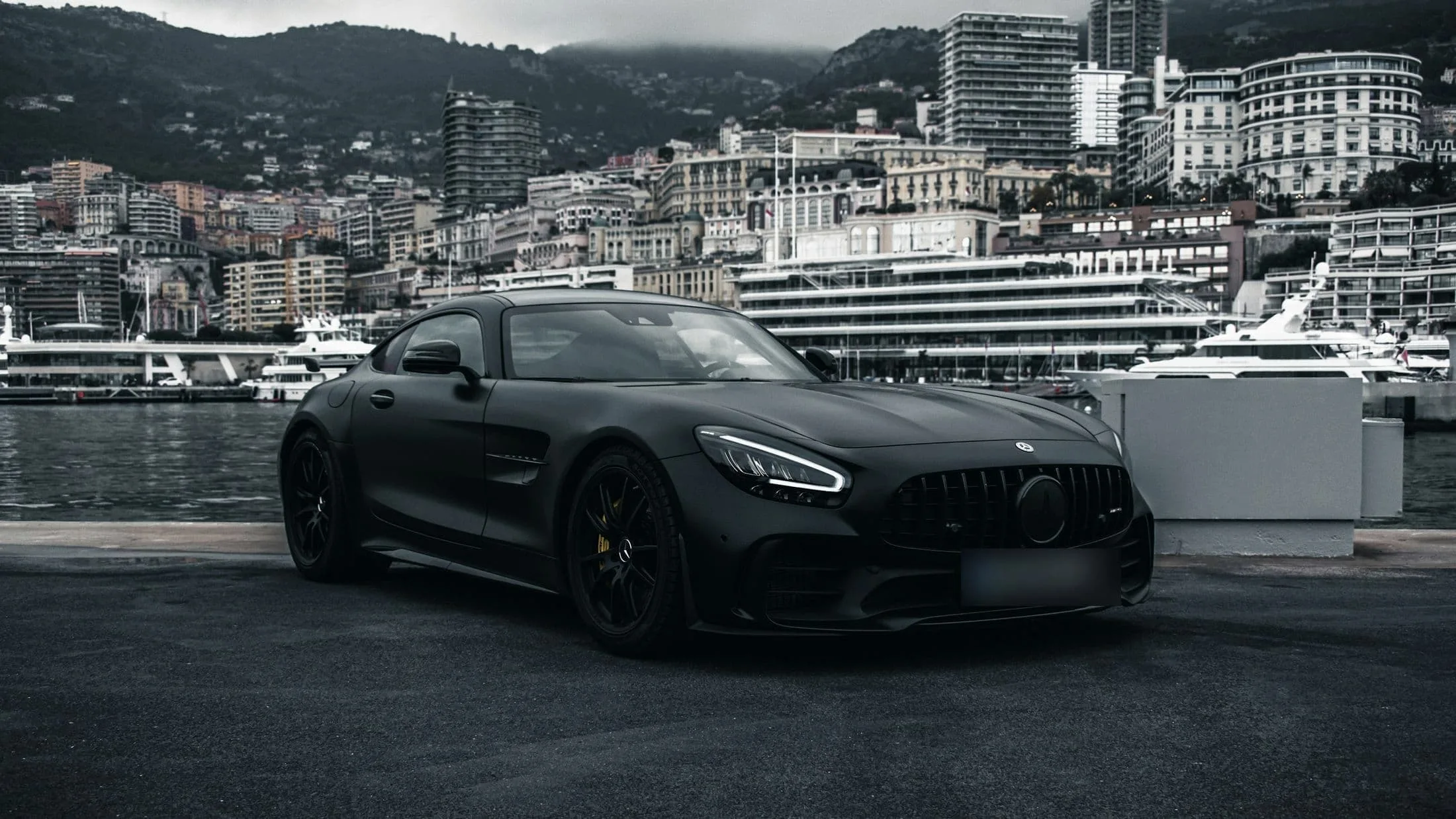 Matte black Mercedes-AMG GT parked on a harbour front with a hillside city behind