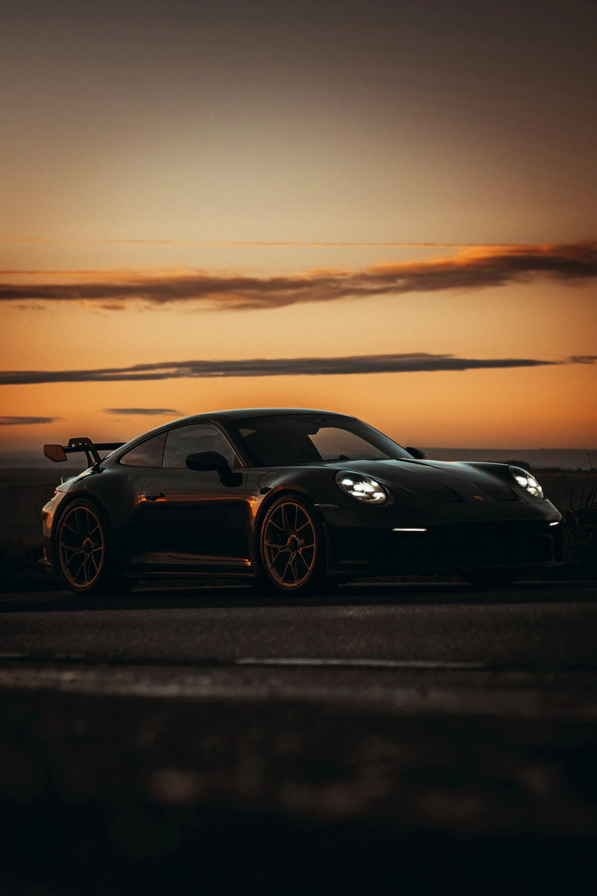 Black Porsche 911 GT3 on an empty coast road at sunset, a band of orange sky above the horizon