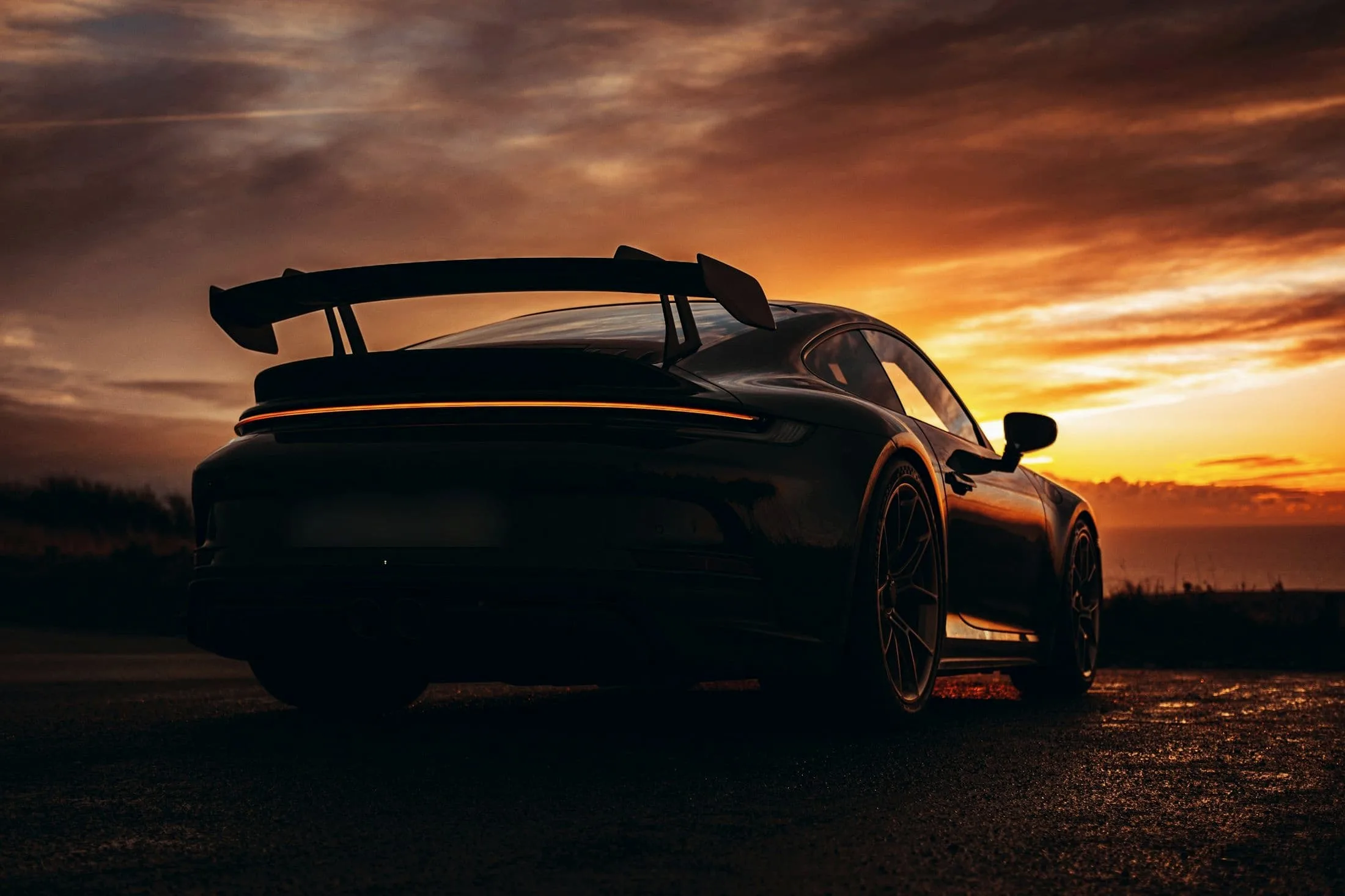 Porsche 911 GT3 rear three-quarter in silhouette against a burning sunset, swan-neck wing raised
